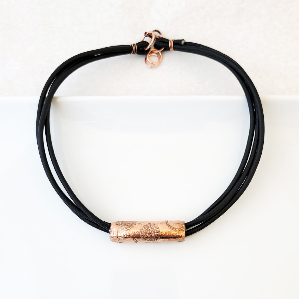 Etched circle design on copper tube on black leather necklace.
